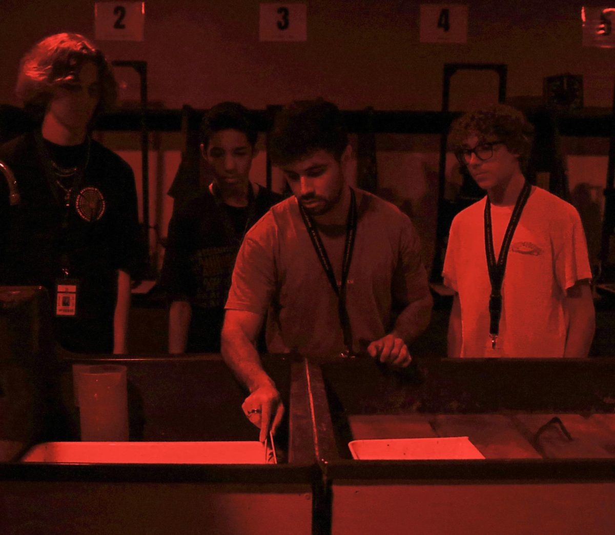 Surrounded by his students, Mr. Angulo demonstrates the process of developing an image in the darkroom. He transferred the paper from the developing chemical to the stop bath using a pair of tongs. To protect the light-sensitive paper in the darkroom, students must leave their phones, watches, and other devices with lights outside.